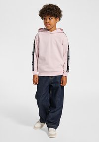 Lichtroze hoodie met een trekkoord en zwarte merkstrepen langs de mouwen, gecombineerd met losse donkere spijkerbroeken en witte sneakers.