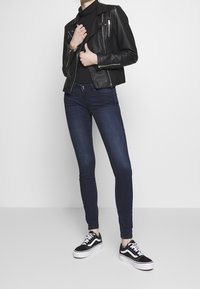 Chaqueta de motociclista de cuero negro sobre un suéter negro ajustado, combinada con jeans oscuros ajustados y zapatillas negras con detalles en blanco.