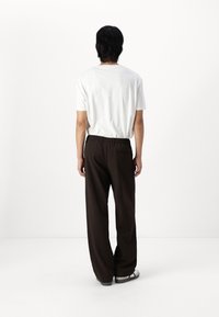 Biała koszula z krótkim rękawem, luźne brązowe spodnie z elastycznym pasem i tylną kieszenią. Czarne sandały z szarą podeszwą. Jednolite tło.