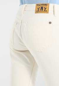 Jeans de denim color crema con un acabado texturizado, que presentan una etiqueta de cuero y bolsillos traseros. Diseño limpio con detalles de hardware mínimos.
