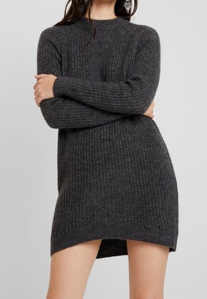 Femme portant une robe-pull en maille côtelée gris foncé à manches longues et des boucles d'oreilles géométriques pendantes, posant devant un fond uni.