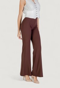 Pantaloni a campana marroni aderenti in un materiale elasticizzato, abbinati a un top in satin bianco aderente con chiusura a bottoni sul davanti.