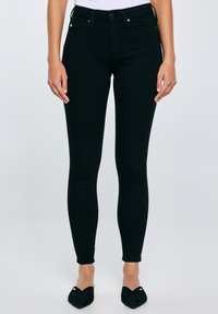 Sieviete melnās saspringtās džinsās un melnos zābaciņos ar noapaļotiem galiem, stāvot pret plānu gaišu fonu.