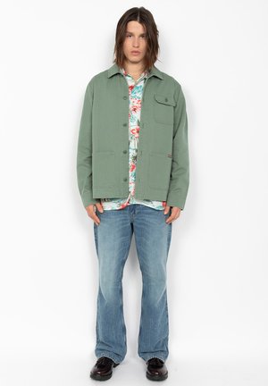 Ung person med skulderlangt hår iført en grøn knapjakke over en blomsterprintet skjorte, løse blå jeans og sorte sko, stående mod hvid baggrund.