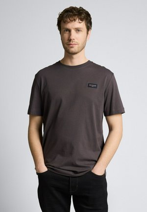 Jonge man met kort krullend haar, draagt een donkergrijs Tom Tailor t-shirt en zwarte broek, handen in de zakken, neutrale achtergrond.