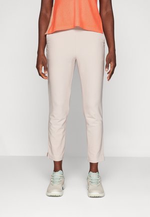 Person iført lyse beige slim-fit bukser, mintgrønne og hvide sneakers og en orange ærmeløs top, stående mod en ensfarvet baggrund.