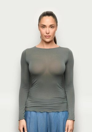 Donna con capelli castani raccolti, che indossa una maglia grigia aderente a maniche lunghe e pantaloni blu larghi, in piedi contro uno sfondo chiaro e semplice.