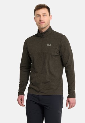 Uomo con un pullover verde scuro con zip sul quarto superiore e pantaloni neri, in piedi e che guarda leggermente verso l'alto e di lato.