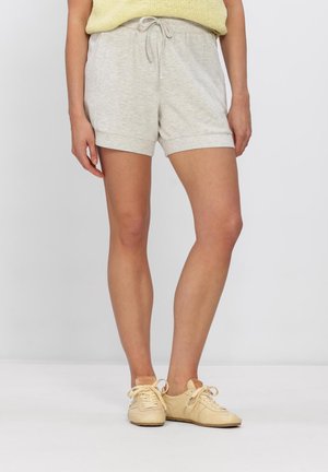 Personne portant un short gris clair à cordon, un haut en maille jaune et des baskets beige à lacets, debout devant un fond blanc uni.