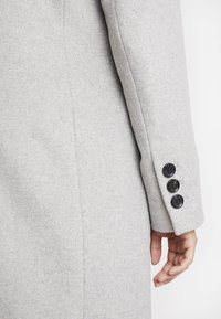 Light grey wool coat sleeve with a structured design, featuring three black buttons and a smooth texture. Hand resting gently on the fabric.
