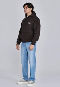 Mörkbrun bomullshoodie med framficka och broderad logotyp, kombinerad med ljusblå jeans i denim och grå sneakers. Enkel, avslappnad passform.