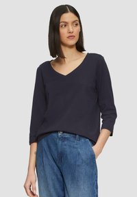 Marineblauw V-hals top gemaakt van zacht materiaal, met driekwart mouwen en een relaxte pasvorm, gecombineerd met lichtblauwe geplooide jeans.