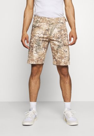 Hombre con pantalones cortos tipo cargo en camuflaje beige y marrón, calcetines blancos, zapatillas blancas y camiseta sin mangas blanca, de pie frente a un fondo liso.