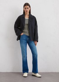 Schwarzer doppelt gefalteter Trenchcoat über einem gestreiften Hemd, kombiniert mit blauen Schlagjeans und weißen Slippern. Schlichtes, strukturiertes Design.