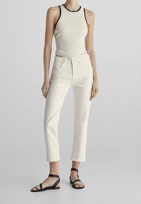 Débardeur crème à côtelés avec des bordures noires, associé à un jean beige clair cropped et des sandales à lanières noires, mettant en valeur une silhouette ajustée.