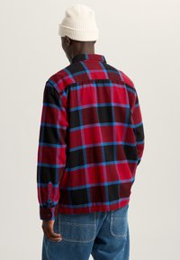 Flanellen shirt in rood en zwart met blauw geruit patroon. Voorzien van lange mouwen, knopenmanchetten en een klassieke kraag. Draagt bij denim broek.