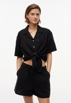 Kvinde iført en sort kortærmet skjorte med knapper, bundet i taljen, og matchende sorte shorts med elastik i taljen, hænder i lommerne.