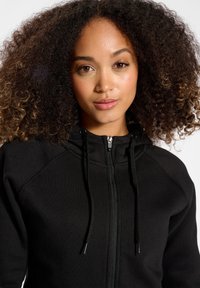 Felpa con zip nera dal tocco morbido, dotata di cappuccio, lacci e polsini a costine. Design semplice senza fantasie o decorazioni.