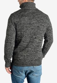 Sweter z dzianiny w ciemnoszarym i czarnym melanżowym wzorze, z wysokim kołnierzem oraz ściągaczami na dolnym końcu i rękawach, w parze z niebieskimi dżinsami.