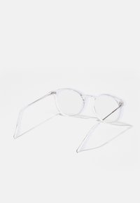 Caurspīdīgas apaļas brilles ar caurspīdīgu plastmasas rāmi un plānām metāla kājām. Lēcas ir caurspīdīgas, bez redzamas krāsas vai raksta.