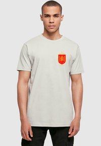 Světle šedé bavlněné tričko s červeným a žlutým státním emblémem Španělska na levé části hrudi. Krátké rukávy a kulatý výstřih.