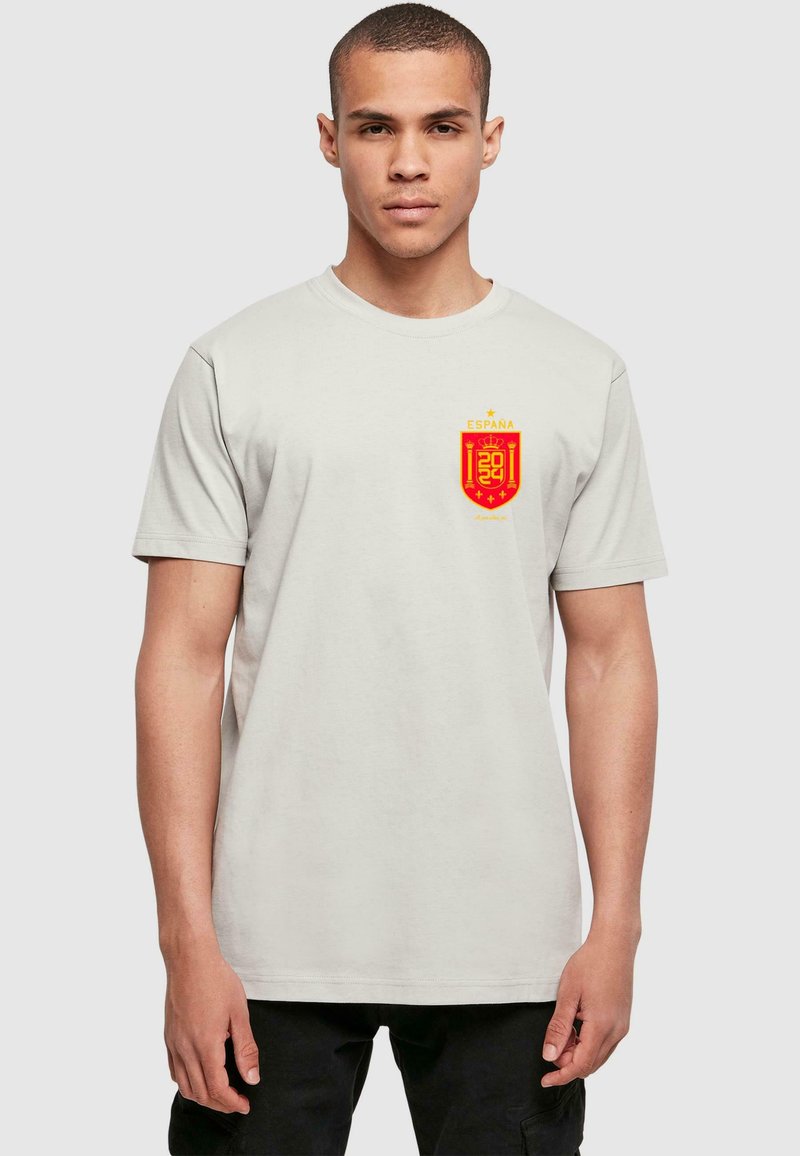 Světle šedé bavlněné tričko s červeným a žlutým státním emblémem Španělska na levé části hrudi. Krátké rukávy a kulatý výstřih.