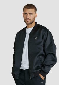 Giacca bomber nera realizzata in materiale liscio, con polsini e colletto a coste, caratterizzata da un piccolo logo sul petto. Indossata sopra una t-shirt bianca.