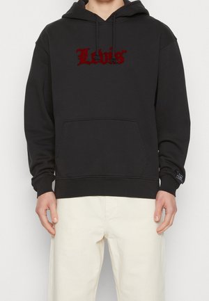 Osoba nosząca czarną bluzę z kapturem z czerwonym logo "Levi's", przednią kieszenią i sznurkami, w połączeniu z kremowymi spodniami.