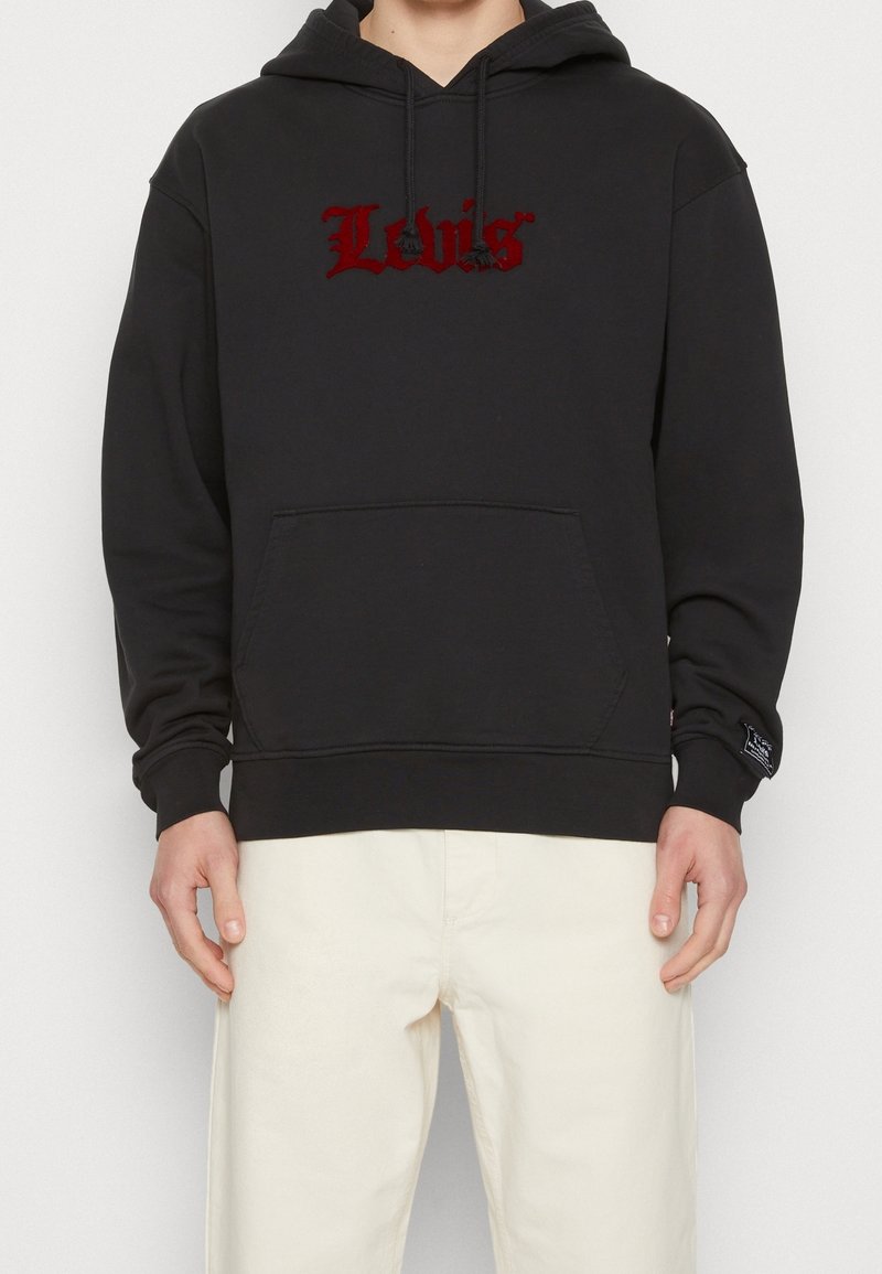 Personne portant un sweat à capuche noir avec le logo rouge "Levi's", une poche avant et des cordons, associé à un pantalon écru.