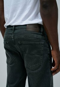 Jeans in denim grigio scuro con una texture liscia, dotati di due tasche posteriori e un'etichetta in pelle marrone con la scritta "SALSA JEANS" impressa.