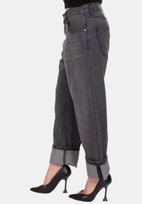 Donna che indossa jeans in denim grigio scuro arrotolati con scarpe con tacco nere, vista di lato su uno sfondo bianco.