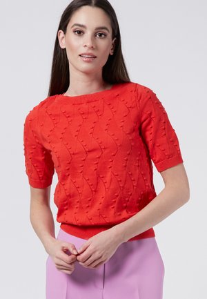 Maglione rosso a maniche corte con colletto a coste e motivo a onda testurizzato. Indossato con un pantalone lilla, caratterizzato da una silhouette aderente.