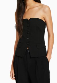 Top nero senza spalline con design aderente, caratterizzato da una chiusura con bottoni sul davanti e due tasche a toppa ai lati. Tessuto liscio e linea sagomata.