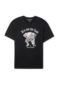 T-shirt nera con vestibilità comoda. Presenta una grafica di mani scheletriche che tengono carte da gioco e testo in bianco: "Non è colpa mia se sono così fortunato."