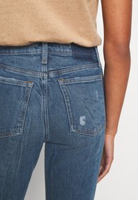 Jeans in denim blu con lavaggio medio, caratterizzati da zone sbiadite, due tasche posteriori e cuciture a contrasto. Il tessuto appare testurizzato.
