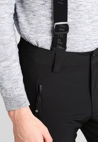 Mustat housut, joissa on säädettävät mustat henkselit, varustettu vetoketjullisilla sivutaskuilla ja sileällä pinnalla, päällä vaaleanharmaa neule.