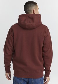 Burgunderfarget hettegenser med stor hette, avslappet passform og ribbestrikkede mansjetter. Laget av et mykt stoff med en glatt tekstur. Bakview synlig.