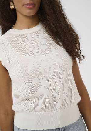 Femme portant un haut en maille sans manches blanc à motifs floraux, une boucle d'oreille créole en or et un collier, avec des cheveux bouclés tombant sur une épaule.