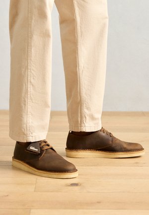 Bruine leren schoenen met een gladde afwerking, ronde neus en lichtgekleurde rubberen zolen. Vergezeld van lichtbeige broek.