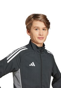 Schwarze Sportjacke mit weißen Akzenten, mit Reißverschluss vorne, hohem Kragen und drei weißen Streifen auf beiden Schultern.