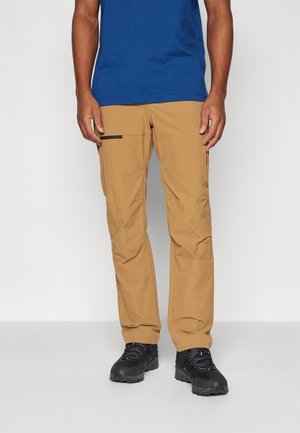 Mann trägt beige Cargohosen mit schwarzen Reißverschlusstaschen, schwarze Wanderschuhe und ein dunkelblaues T-Shirt, steht vor einem einfarbigen Hintergrund.