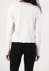 Vit långärmad tröja med ribbad textur och delikat mönster, med en scalloped nederkant, i kombination med svarta leggings.