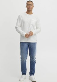 Langærmet, hvid struktureret skjorte med en brystlomme, kombineret med lyseblå jeans og hvide sneakers, sat mod en ensfarvet baggrund.