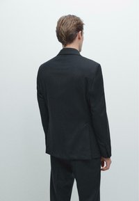 Giacca da suit nera slim-fit, realizzata in tessuto testurizzato, con colletto a forma di scollatura, spalline sottili e dettagli con bottoni sui polsini.
