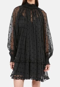 Schwarzes Kleid aus transparenter Stoff mit Polka-Dots, ausgestattet mit einem hohen gerafften Kragen, langen Ärmeln und Rüschen am Saum.