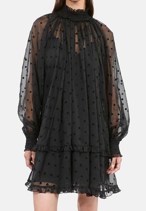 Vestido negro de tela transparente con lunares, con un cuello smock alto, mangas largas y detalles de dobladillo con volantes.