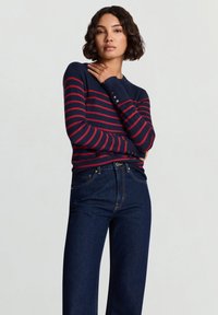 Top a maniche lunghe a righe blu navy e rosse con scollo rotondo e dettagli con bottoni, abbinato a jeans a vita alta in denim scuro.