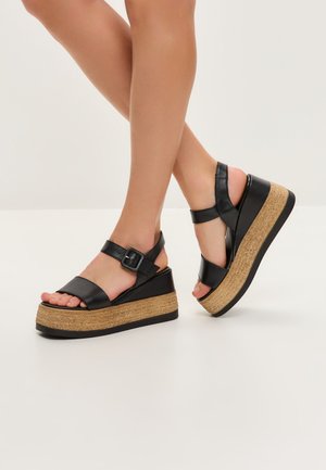 Sandalias de cuña de piel negra con plataforma de corcho, correa ajustable en el tobillo, diseño de punta abierta y superficie texturizada en la suela.
