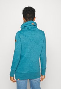 Sudadera color azul verdoso con un cuello alto y detalles de costuras diagonales. Presenta puños y dobladillo acanalados, parche con logo en el cuello y un material suave.