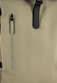 Zaino impermeabile beige con ferramenta nera, dotato di una tasca frontale con zip e tracolla regolabile. Texture liscia, design minimalista.